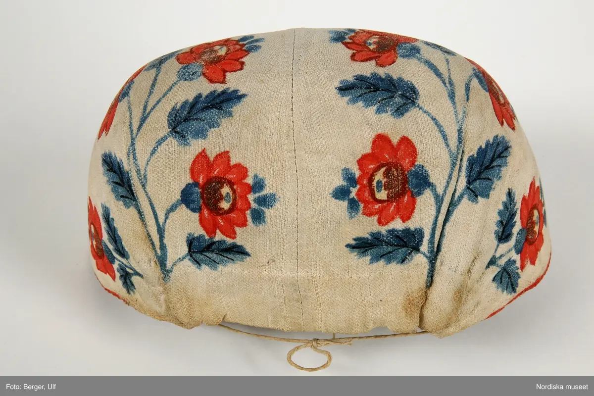 Huvudliggaren bilaga= "Två hvardagsmössor" från Härja socken i Vestergötland, såsom de allmänt begagnades på 1830-talet af gifta qvinnor."

a. Bindmössa, mått från öra till öra 35 cm, djup 11 cm.
b. Bindmössa längd från öra till öra 37 cm

a. av vitt stärkt bomullstyg i panama sydd över en stomme av grå lumppapp, foder saknas. Blommönster i rött och blåttoch något gult  är schablonmålat på tyget efter det att tyget är monterat.Tunn målad röd kant. Tunt dragsnöre i nacken.

b. lik a. av tyg målad i blått med 2 rader blommotiv i rött och grönt och något svart, inuti märkt M med bläck.Knytband i nacken.


Berit Eldvik mars-2005