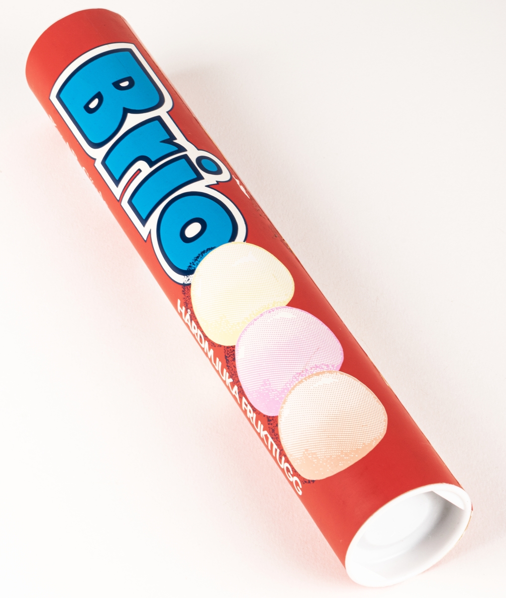 Pappersförpackning utan innehåll. Rör med vita plastlock i var ände. Text i blått och vitt. "BRIO, hårdmjuka frukttugg, Nyhet!"