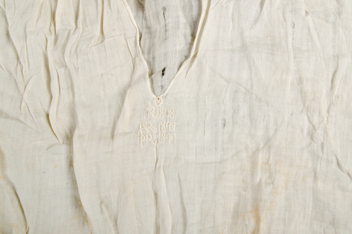 Skjorta för barn. Vit bomullslärft. Långa ärmar med trekantskil. Vit spets på kragen och vid handlederna. Sprund framtill utan knapper. Små knytband av vitt siden vid handlederna. Nedanför sprundet broderat i vitt initialer och årtal: R 1808, IAR 1844 P-R 1847.
Små hål här och var på både framsidan , ärmarna och baksidan. tyget är mer trasigt på skjortans baksida. Stort hål under högra ärmen. Fläckar på baksidan upptill, framsidan nedtill m.m.