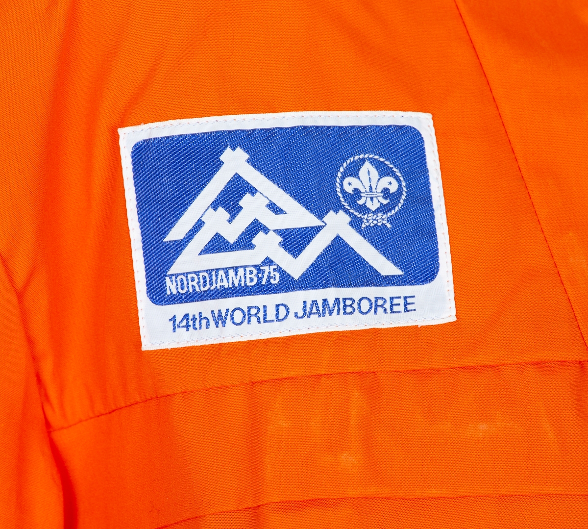 Anorakk. Hette. Brystlomme. Påsydd blått og hvitt merke  merke: Norjamb 75 14. woeld jamboree. menber made of Swiss Polyester
Givers ektemann Per Ottesen var disponent for Speidersport og hadde leveranser av utstyr og merkeklær til deltakere på leiren