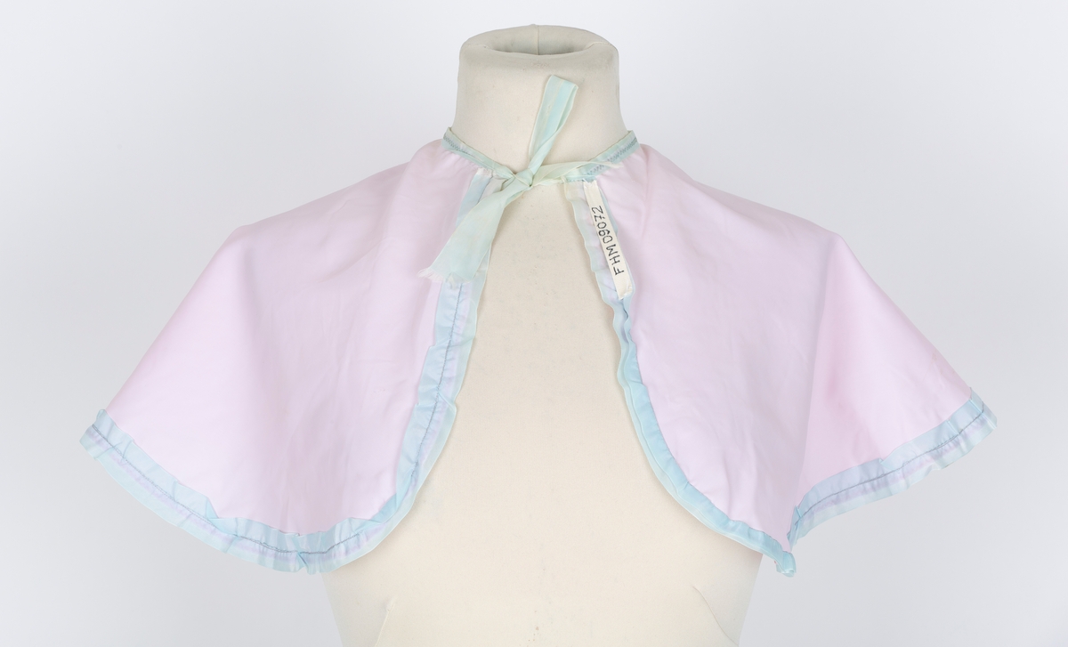 Friserkappe, halvcirkel, avrundet med åpning i en side. Knytebånd. Rosa nylon, lys blå kantrysj.
Brukt over finklær i mange år for å unga flassing på klærne.