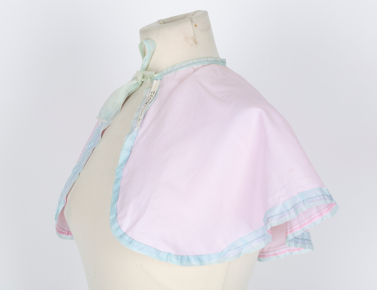 Friserkappe, halvcirkel, avrundet med åpning i en side. Knytebånd. Rosa nylon, lys blå kantrysj.
Brukt over finklær i mange år for å unga flassing på klærne.