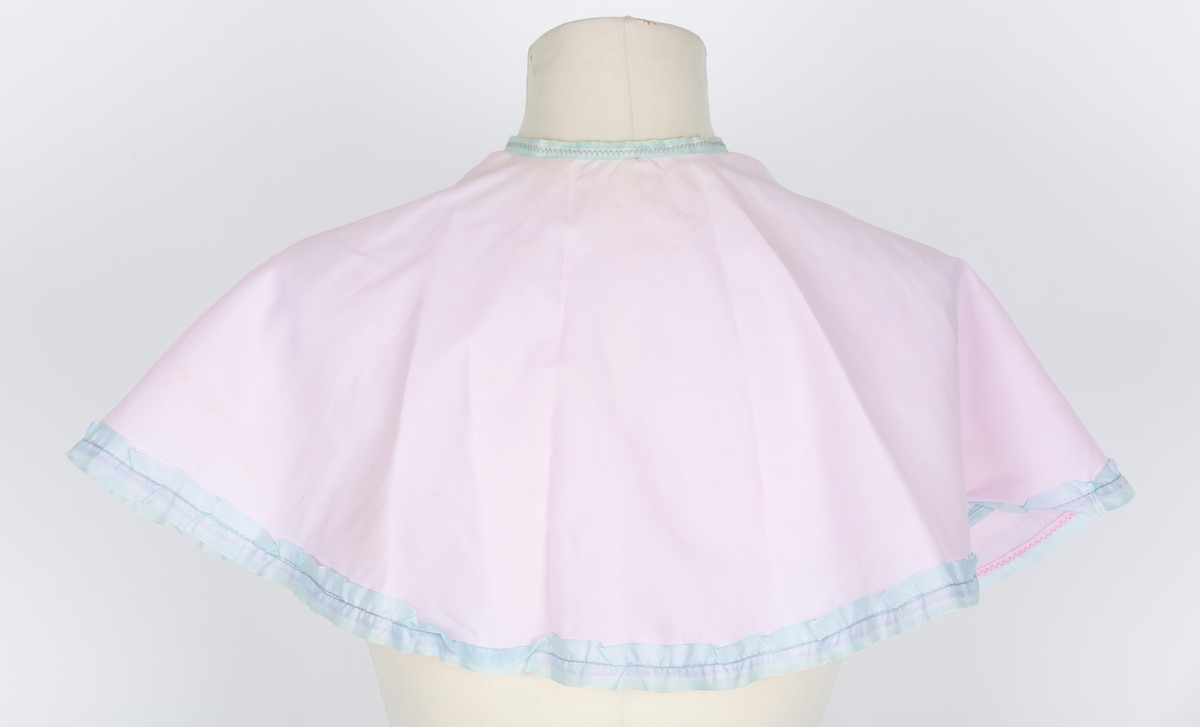 Friserkappe, halvcirkel, avrundet med åpning i en side. Knytebånd. Rosa nylon, lys blå kantrysj.
Brukt over finklær i mange år for å unga flassing på klærne.