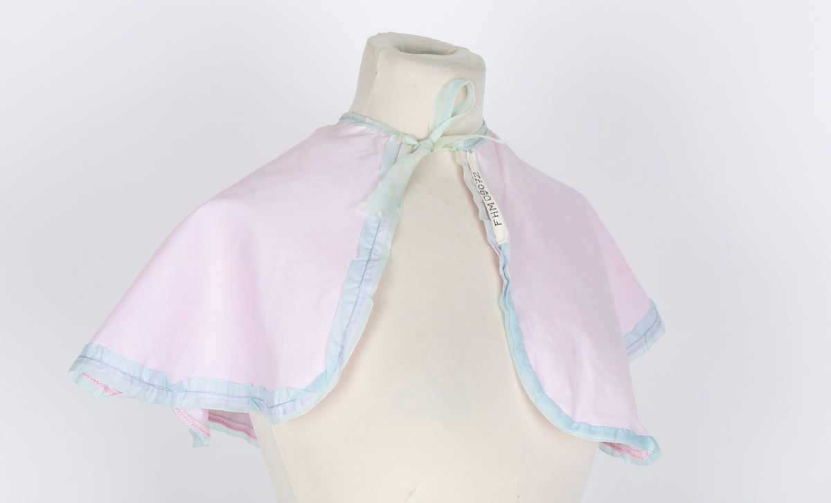 Friserkappe, halvcirkel, avrundet med åpning i en side. Knytebånd. Rosa nylon, lys blå kantrysj.
Brukt over finklær i mange år for å unga flassing på klærne.