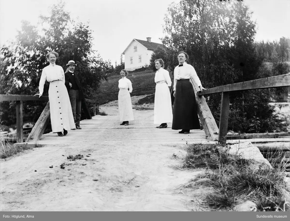 Fyra kvinnor och en man står på en bro över Fanbyån, Stöde, med ett bostadshus i bakgrunden, fotograferat österut mot Råvlan, Östanå.