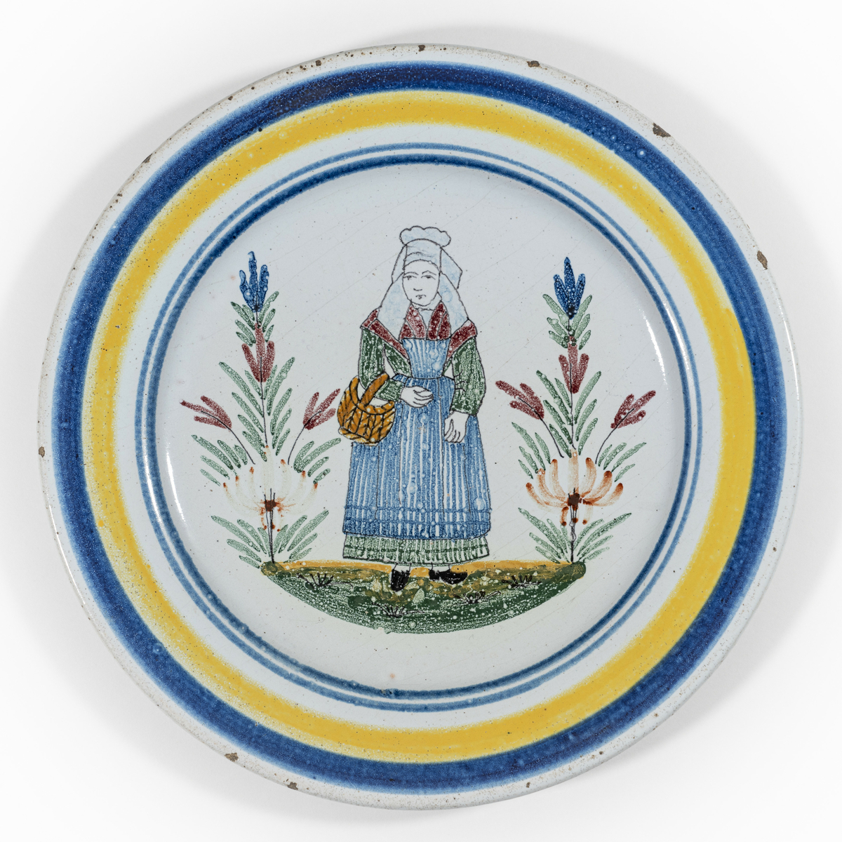 Tallerken i keramikk. Påmalt dekor med kvinne i tradisjonell drakt (stakk, forkle, sjal, spesielt hodeplagg) som holder en kurv. Blomsterranker på hver side av kvinnen. Kant malt med blå og gule ringer. Quimperware. Har tilhørt Sara Fabricius.
