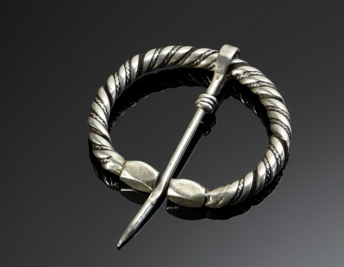 Vikingatida ringspänne av silver. Utgörs av en vriden ring med rektangulära men avfasade ändknoppar. Kvarsittande och lös torne tillverkad av en trind silverten som böjts runt ringspännet och avslutats genom att viras tre varv runt tornen. Ringspännets dekor skapas av att en bredare slät och en tunnare vriden tråd har vridits tillsammans. Dekoren är sliten av användning och på något enstaka ställe har den tunna, vridna tråden lossnat, men i övrigt komplett.
