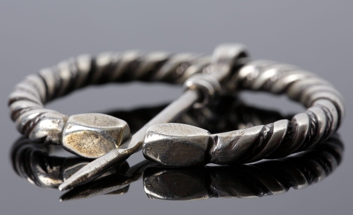 Vikingatida ringspänne av silver. Utgörs av en vriden ring med rektangulära men avfasade ändknoppar. Kvarsittande och lös torne tillverkad av en trind silverten som böjts runt ringspännet och avslutats genom att viras tre varv runt tornen. Ringspännets dekor skapas av att en bredare slät och en tunnare vriden tråd har vridits tillsammans. Dekoren är sliten av användning och på något enstaka ställe har den tunna, vridna tråden lossnat, men i övrigt komplett.