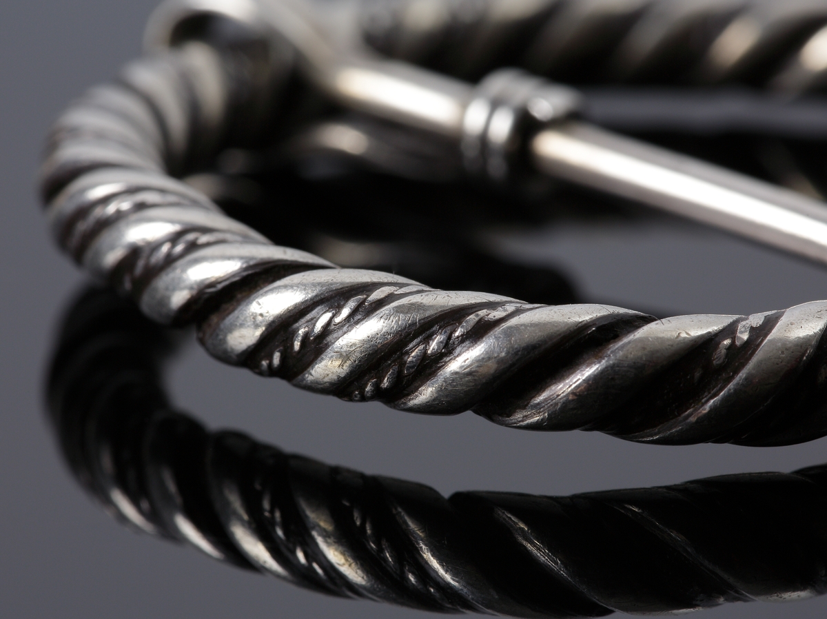 Vikingatida ringspänne av silver. Utgörs av en vriden ring med rektangulära men avfasade ändknoppar. Kvarsittande och lös torne tillverkad av en trind silverten som böjts runt ringspännet och avslutats genom att viras tre varv runt tornen. Ringspännets dekor skapas av att en bredare slät och en tunnare vriden tråd har vridits tillsammans. Dekoren är sliten av användning och på något enstaka ställe har den tunna, vridna tråden lossnat, men i övrigt komplett.