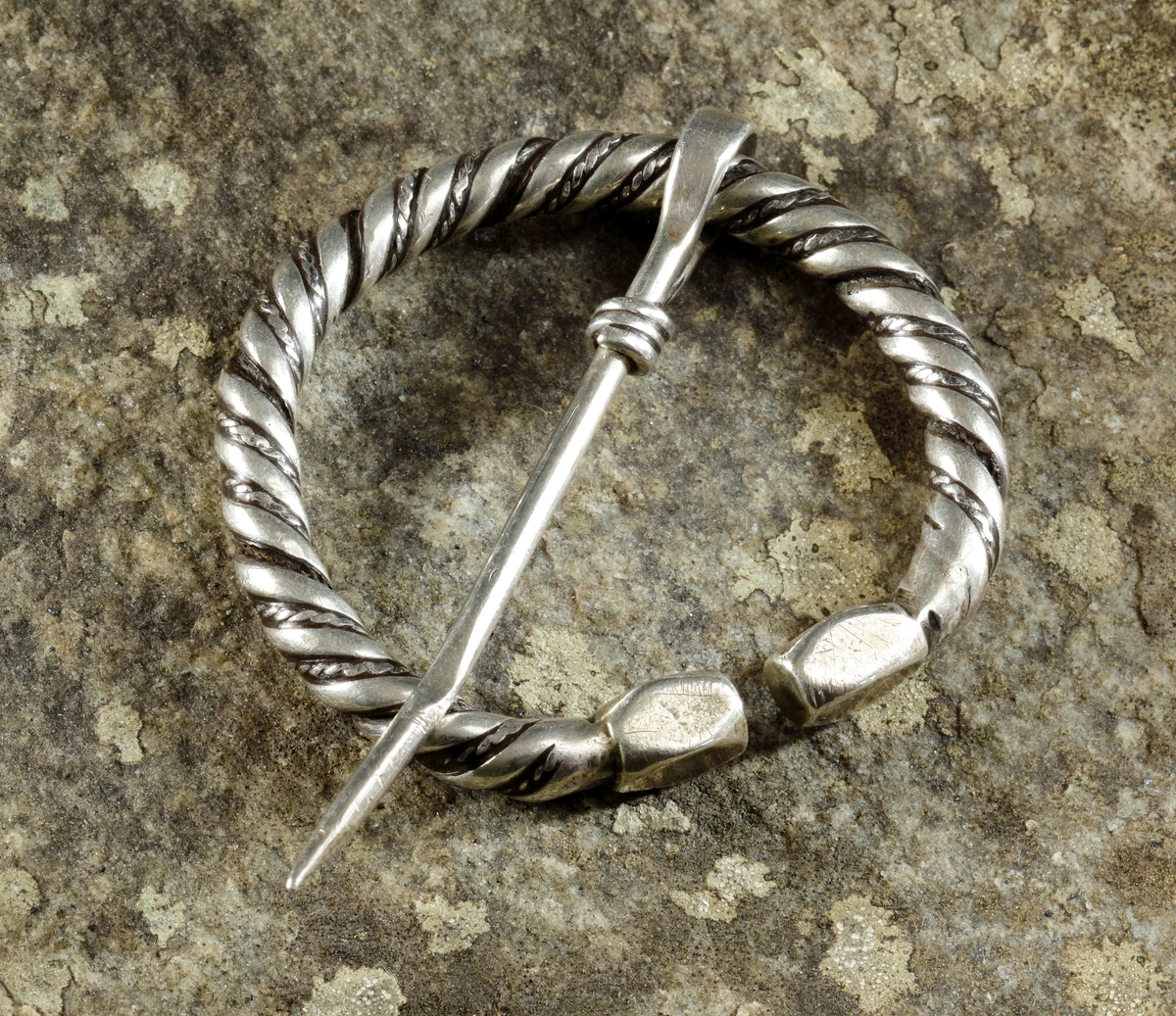 Vikingatida ringspänne av silver. Utgörs av en vriden ring med rektangulära men avfasade ändknoppar. Kvarsittande och lös torne tillverkad av en trind silverten som böjts runt ringspännet och avslutats genom att viras tre varv runt tornen. Ringspännets dekor skapas av att en bredare slät och en tunnare vriden tråd har vridits tillsammans. Dekoren är sliten av användning och på något enstaka ställe har den tunna, vridna tråden lossnat, men i övrigt komplett.
