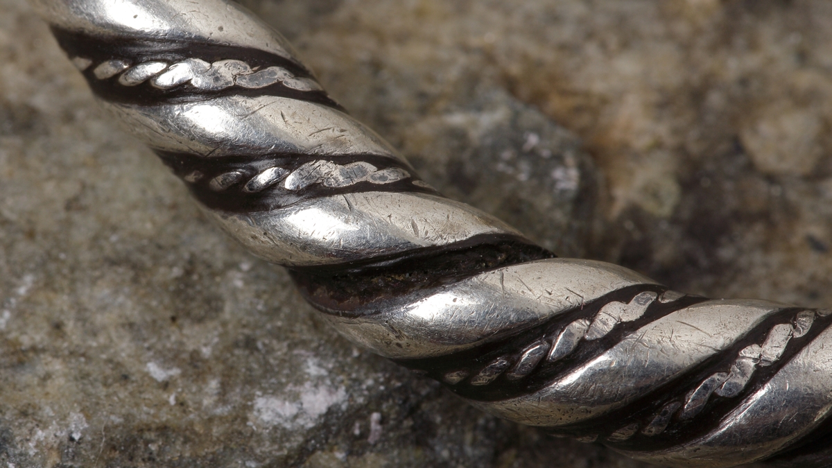 Vikingatida ringspänne av silver. Utgörs av en vriden ring med rektangulära men avfasade ändknoppar. Kvarsittande och lös torne tillverkad av en trind silverten som böjts runt ringspännet och avslutats genom att viras tre varv runt tornen. Ringspännets dekor skapas av att en bredare slät och en tunnare vriden tråd har vridits tillsammans. Dekoren är sliten av användning och på något enstaka ställe har den tunna, vridna tråden lossnat, men i övrigt komplett.