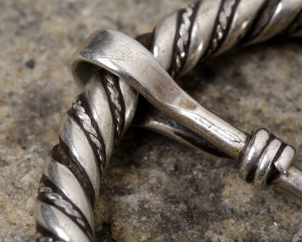 Vikingatida ringspänne av silver. Utgörs av en vriden ring med rektangulära men avfasade ändknoppar. Kvarsittande och lös torne tillverkad av en trind silverten som böjts runt ringspännet och avslutats genom att viras tre varv runt tornen. Ringspännets dekor skapas av att en bredare slät och en tunnare vriden tråd har vridits tillsammans. Dekoren är sliten av användning och på något enstaka ställe har den tunna, vridna tråden lossnat, men i övrigt komplett.