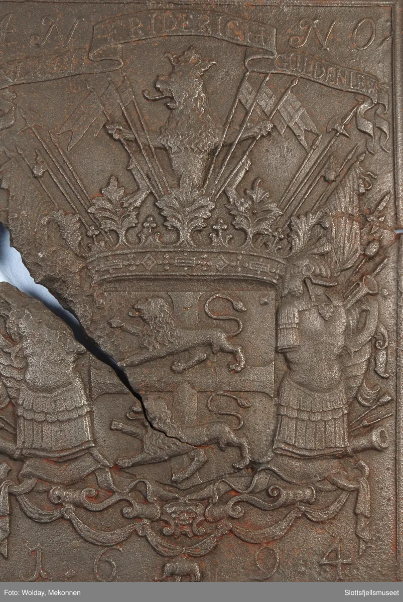 Gyldenløves våpern, omgitt av krigerske emblemer, derover på knekket bånd "ULRICH FRIDERICH"
nederst 1694