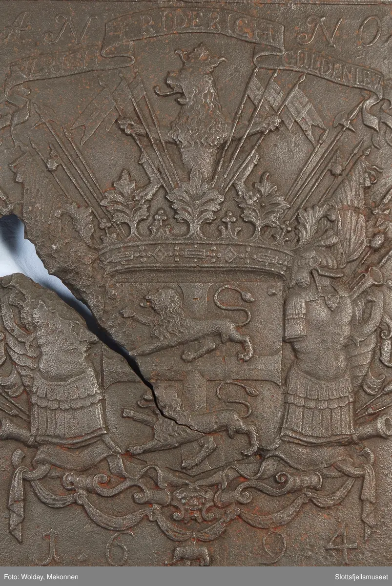 Gyldenløves våpern, omgitt av krigerske emblemer, derover på knekket bånd "ULRICH FRIDERICH"
nederst 1694