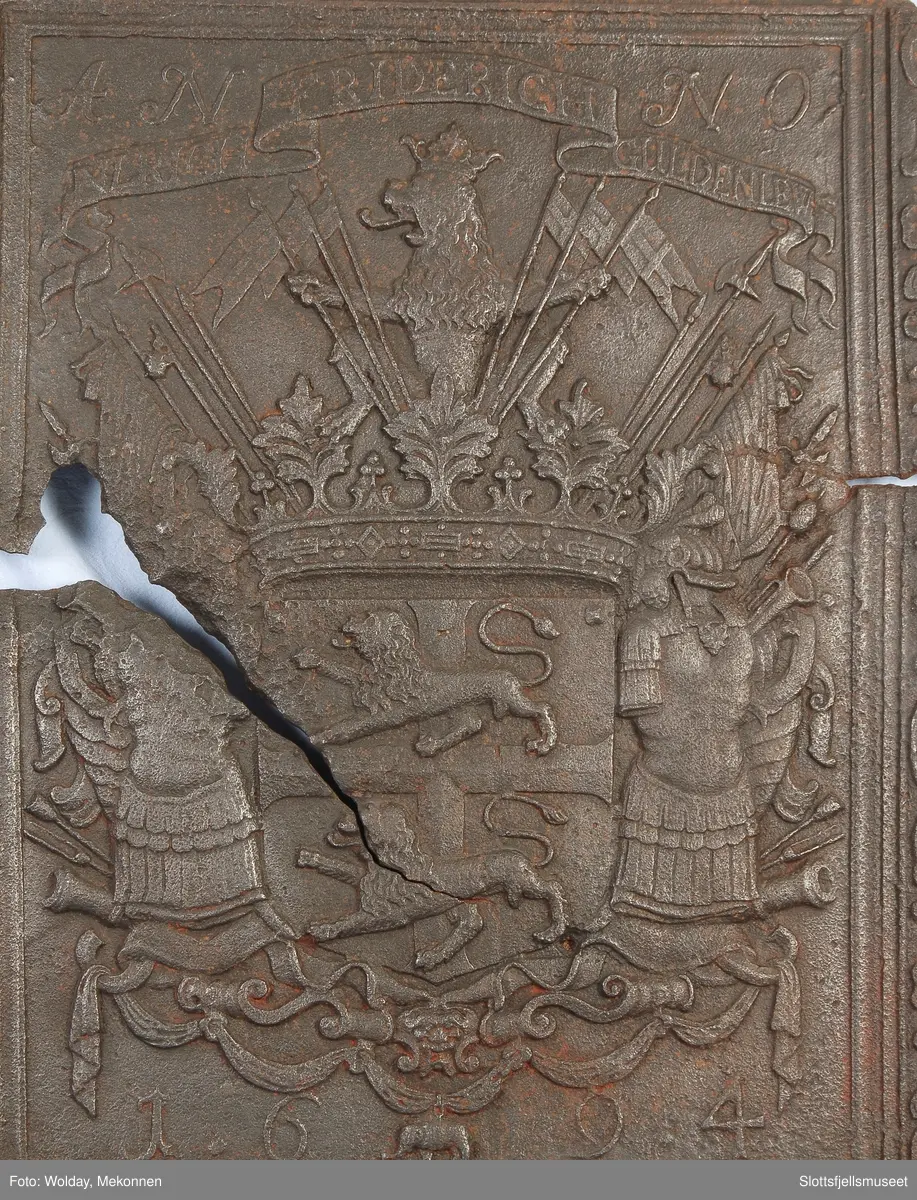Gyldenløves våpern, omgitt av krigerske emblemer, derover på knekket bånd "ULRICH FRIDERICH"
nederst 1694
