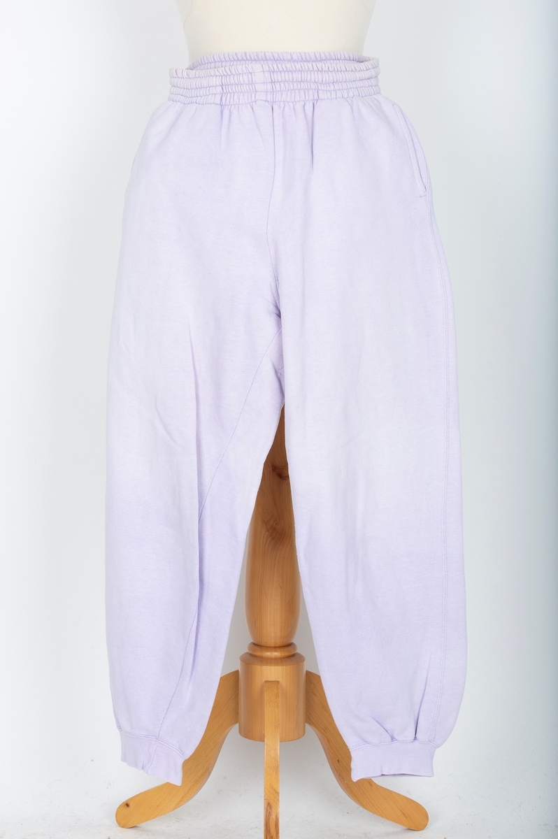 Joggedress. Todelt. Tricot. Lys lilla. Genser med raglan ermer, vrangborder. Bukse med strikk i livet, vrangbord på bukseben.
Brukt til uke-trim og fritid sent 1980-tall