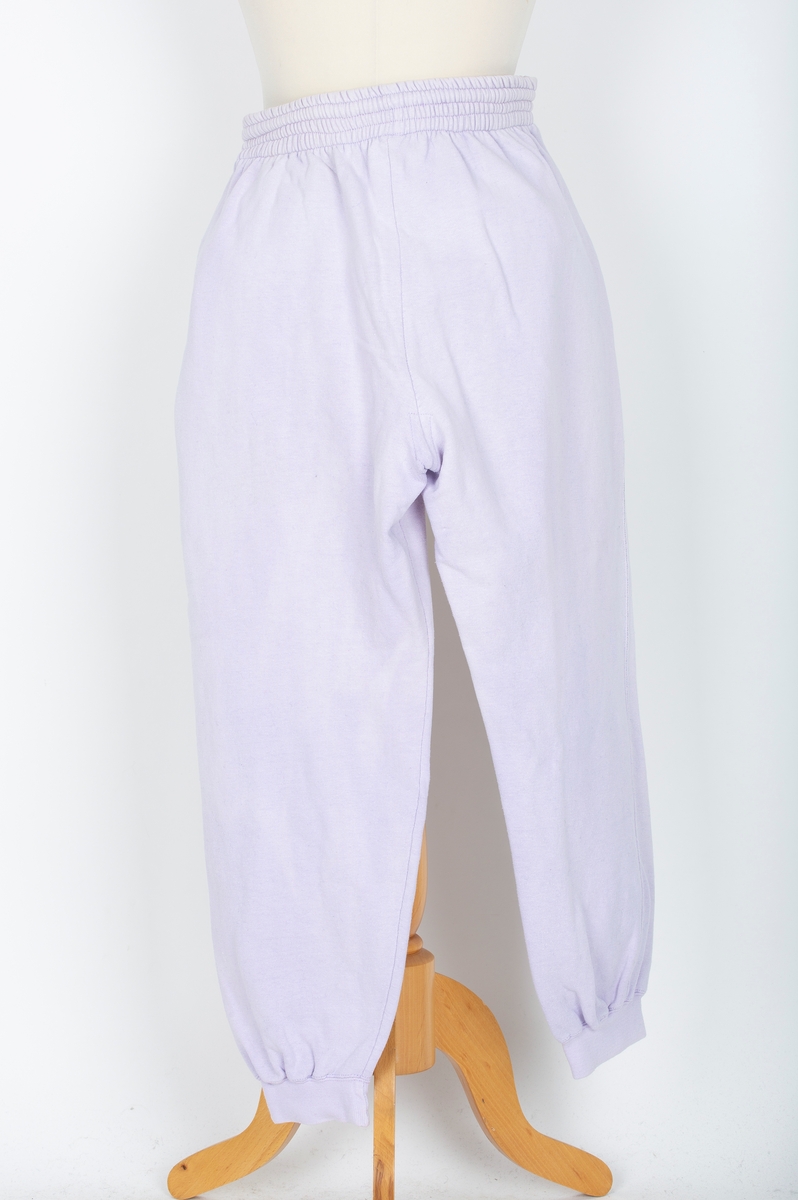 Joggedress. Todelt. Tricot. Lys lilla. Genser med raglan ermer, vrangborder. Bukse med strikk i livet, vrangbord på bukseben.
Brukt til uke-trim og fritid sent 1980-tall
