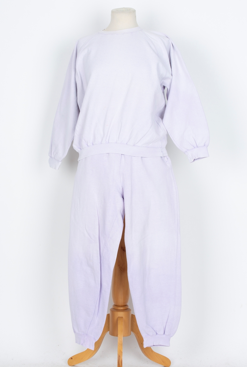 Joggedress. Todelt. Tricot. Lys lilla. Genser med raglan ermer, vrangborder. Bukse med strikk i livet, vrangbord på bukseben.
Brukt til uke-trim og fritid sent 1980-tall