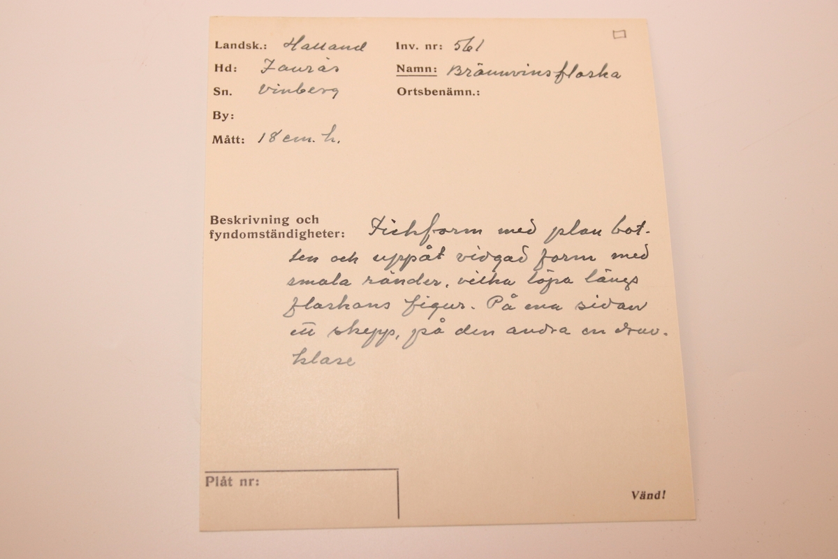 Glasflaska med plan botten och uppåt vidgad form. Med reliefdekor föreställande smala ränder som täcker flaskans ytor. På flaskans ena sidan är ett skepp avbildat, på den andra en druvklase. Med kork.