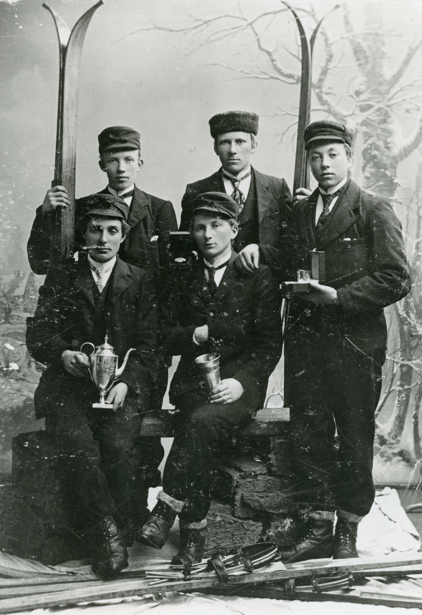 5 yngre menn med ski og premiar fotograferte i atelier. Bildet er teke i etterkant av eit landsrenn i Kikutbakken i Skien i 1904. 
Framme frå v. sit: Olav A. Tjønna og Sveinung G. Bø.
Bak frå v. stend: Olav O. Ajer, Gregar G. Bø og Sveinung G. Kåsa. 
Sveinung G. Bø sit med kongepokalen.