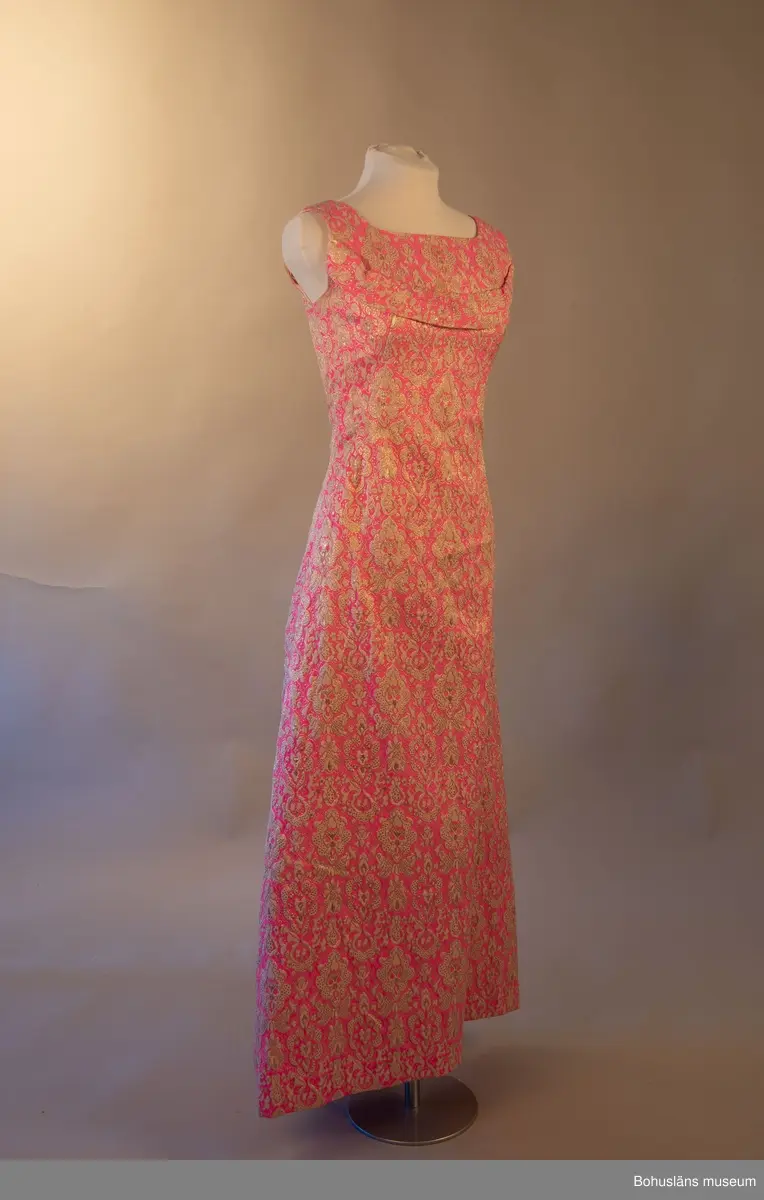 Klänning med beigt och guldfärgat mönster mot rosa botten. Rosett baktill som för blicken fäster det draperade tyget mellan skuldrorna. Framsida utsmyckad med en bård över bysten.