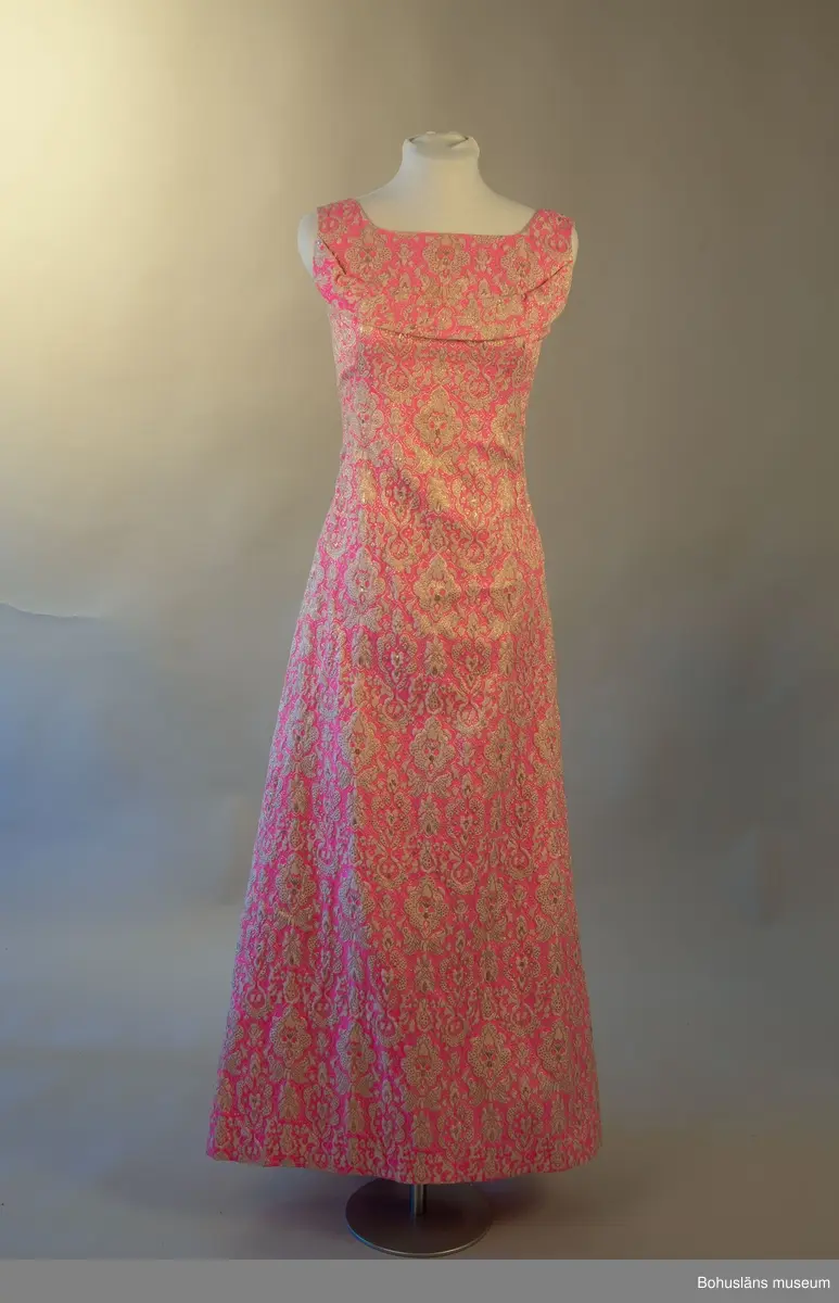 Klänning med beigt och guldfärgat mönster mot rosa botten. Rosett baktill som för blicken fäster det draperade tyget mellan skuldrorna. Framsida utsmyckad med en bård över bysten.