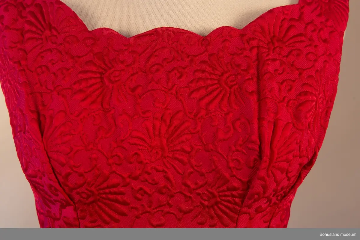 Långklänning i djupröd färg. Tyget är mussel- och girlandornamenterad. Klänningen draperad för att skapa dess fall. Baktill en rosett som samlar upp tyg och skapar klänningens figur. Insida lapp märkt ”Lilbi Modell”.