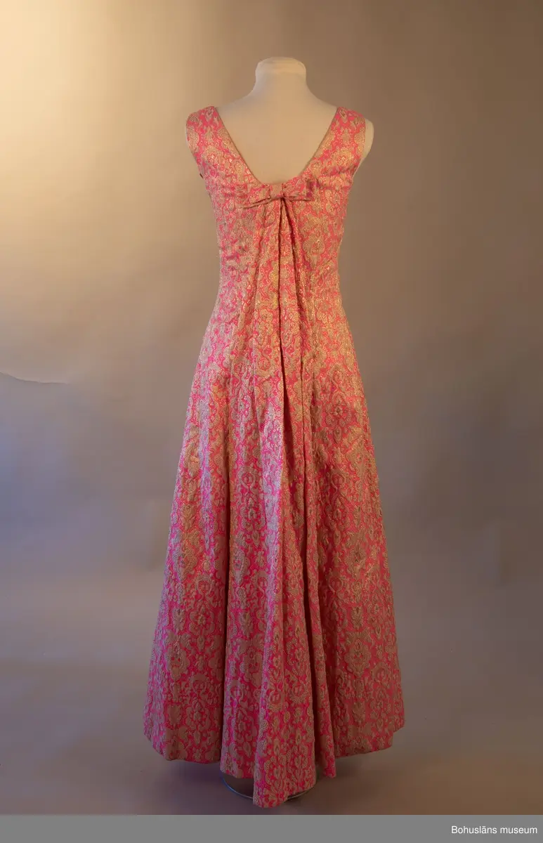 Klänning med beigt och guldfärgat mönster mot rosa botten. Rosett baktill som för blicken fäster det draperade tyget mellan skuldrorna. Framsida utsmyckad med en bård över bysten.