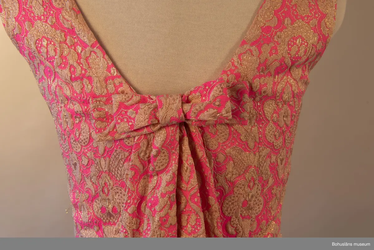 Klänning med beigt och guldfärgat mönster mot rosa botten. Rosett baktill som för blicken fäster det draperade tyget mellan skuldrorna. Framsida utsmyckad med en bård över bysten.