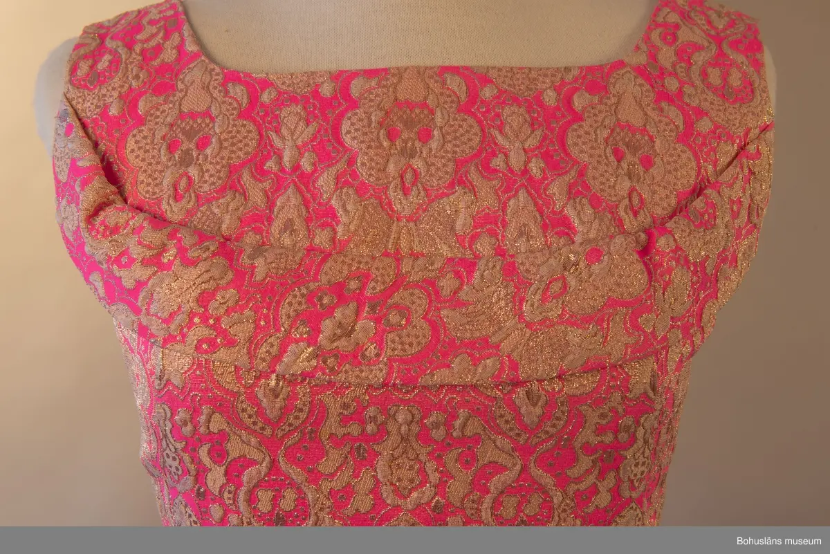 Klänning med beigt och guldfärgat mönster mot rosa botten. Rosett baktill som för blicken fäster det draperade tyget mellan skuldrorna. Framsida utsmyckad med en bård över bysten.