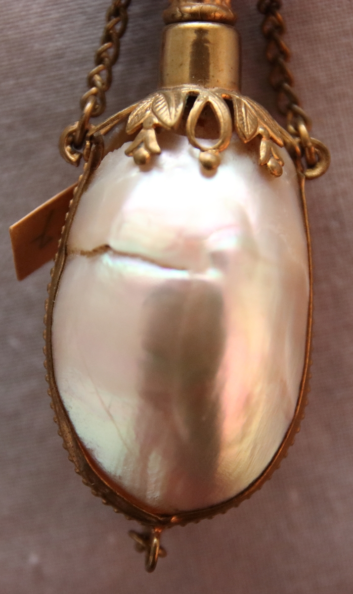 Luktdosa gjord av pärlemoskimrande, ovala snäckskal som hålls samman av en smal ornerad mässingsskena i form av pärlstav. Upptill avslutas dosan av en hals (i formpressad metall i form av blad och bär) i vilken det sitter en kork med ett hölje i mässing. Dosan hänger i en kedja som är fäst i två ringar på sidan av korken och som avslutas med en ring som är dekorerad med en pärlstav. På botten av dosan hänger ett litet droppformat avslut i metallskenan.