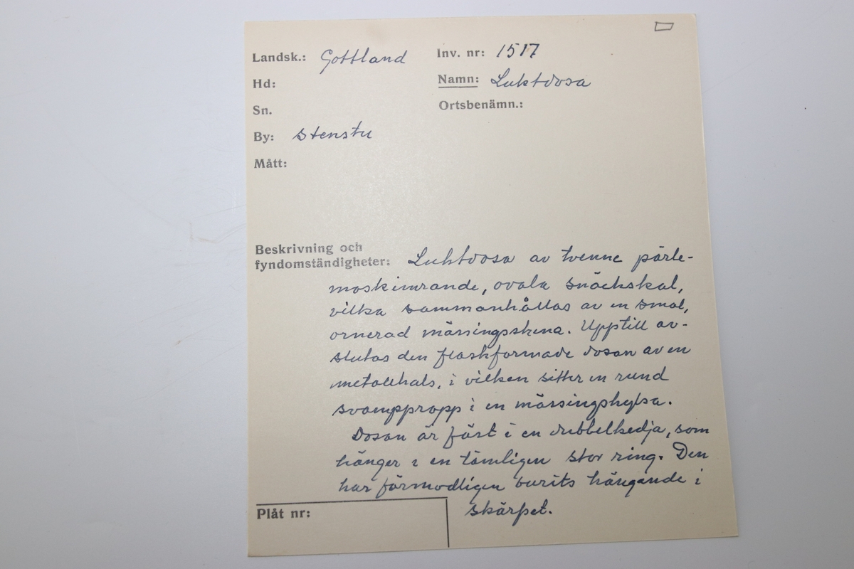 Luktdosa gjord av pärlemoskimrande, ovala snäckskal som hålls samman av en smal ornerad mässingsskena i form av pärlstav. Upptill avslutas dosan av en hals (i formpressad metall i form av blad och bär) i vilken det sitter en kork med ett hölje i mässing. Dosan hänger i en kedja som är fäst i två ringar på sidan av korken och som avslutas med en ring som är dekorerad med en pärlstav. På botten av dosan hänger ett litet droppformat avslut i metallskenan.