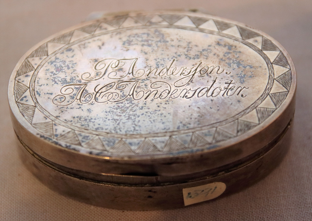 Oval ask i silver. Namnen "P Andersson" och "AC Andersdoter" är inristade på lockets ovansida, omgivna av en dekorativ bård med triagulära mönster. Asken är stämplad på botten på insdian av asken med kattfot, i4 (år 1839) och två svårtydda stämplar.