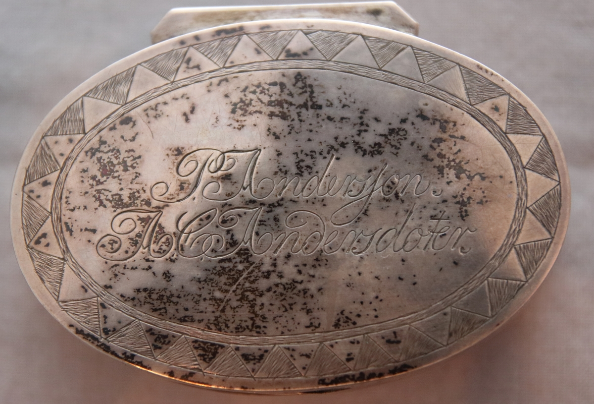 Oval ask i silver. Namnen "P Andersson" och "AC Andersdoter" är inristade på lockets ovansida, omgivna av en dekorativ bård med triagulära mönster. Asken är stämplad på botten på insdian av asken med kattfot, i4 (år 1839) och två svårtydda stämplar.