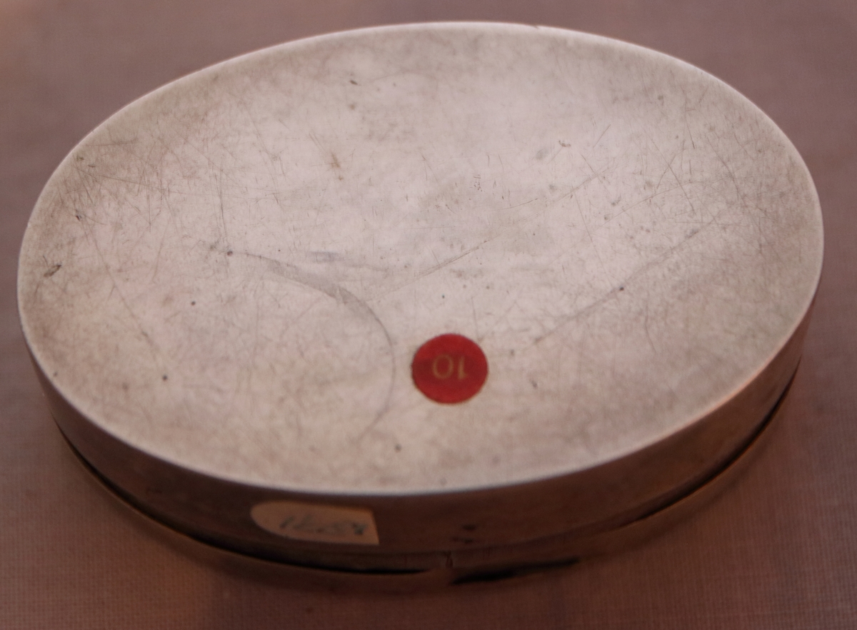 Oval ask i silver. Namnen "P Andersson" och "AC Andersdoter" är inristade på lockets ovansida, omgivna av en dekorativ bård med triagulära mönster. Asken är stämplad på botten på insdian av asken med kattfot, i4 (år 1839) och två svårtydda stämplar.