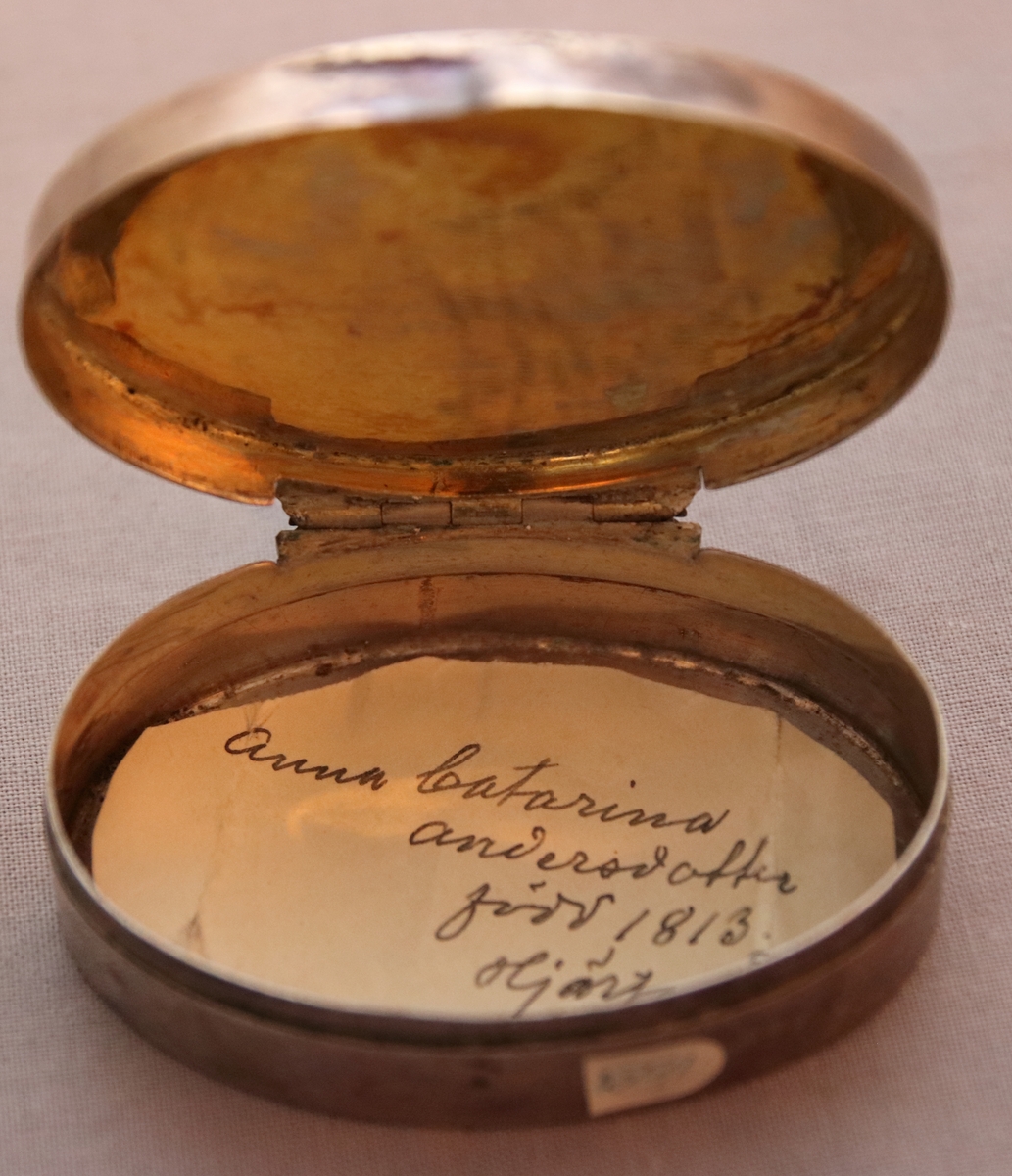 Oval ask i silver. Namnen "P Andersson" och "AC Andersdoter" är inristade på lockets ovansida, omgivna av en dekorativ bård med triagulära mönster. Asken är stämplad på botten på insdian av asken med kattfot, i4 (år 1839) och två svårtydda stämplar.