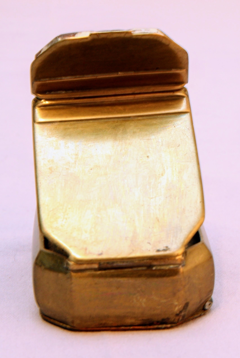 Tobaksdosa i mässing med uppfällbar spegel. Asken öppnas med ett lock i botten. Med gångjärn i ena hörnet där stödet till den uppfällbara spegeln är gängat.  Inskription i botten med bokstäverna ”APL" och siffrorna 1873 efter. Förutom inskriptionen finns inga dekorativa element på föremålet.