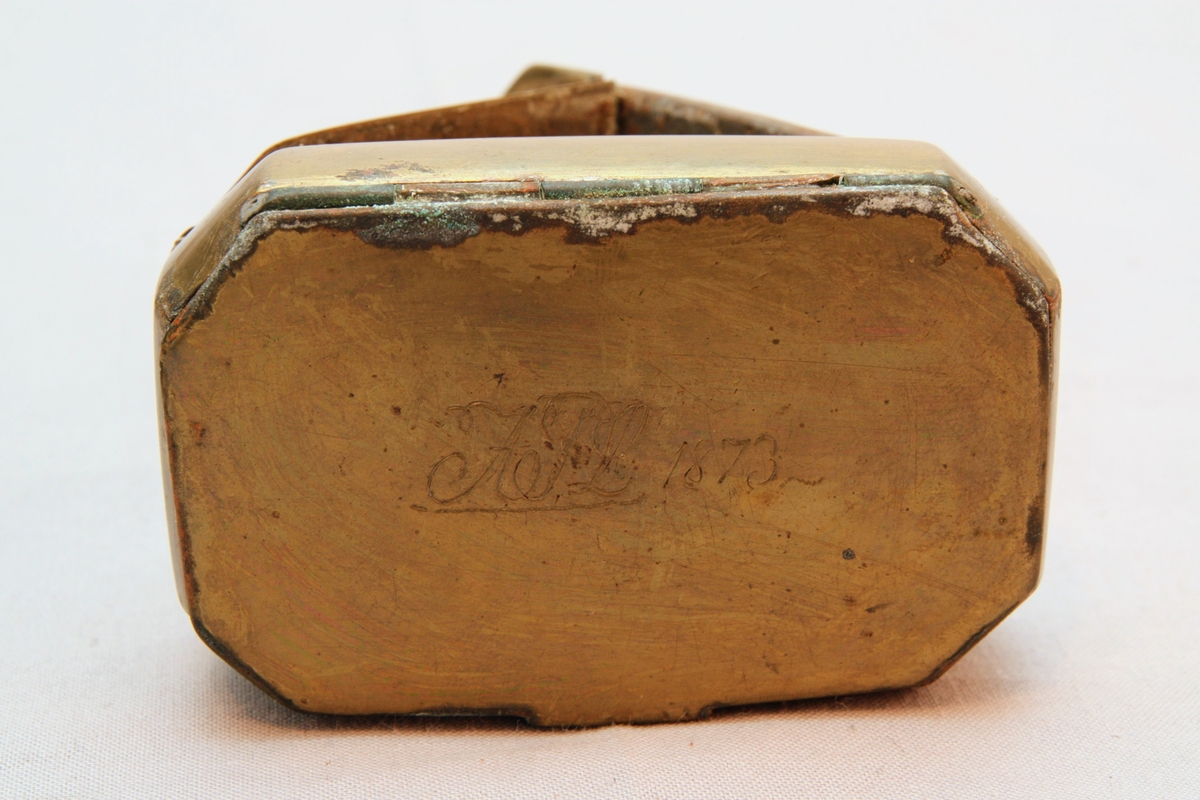 Tobaksdosa i mässing med uppfällbar spegel. Asken öppnas med ett lock i botten. Med gångjärn i ena hörnet där stödet till den uppfällbara spegeln är gängat.  Inskription i botten med bokstäverna ”APL" och siffrorna 1873 efter. Förutom inskriptionen finns inga dekorativa element på föremålet.