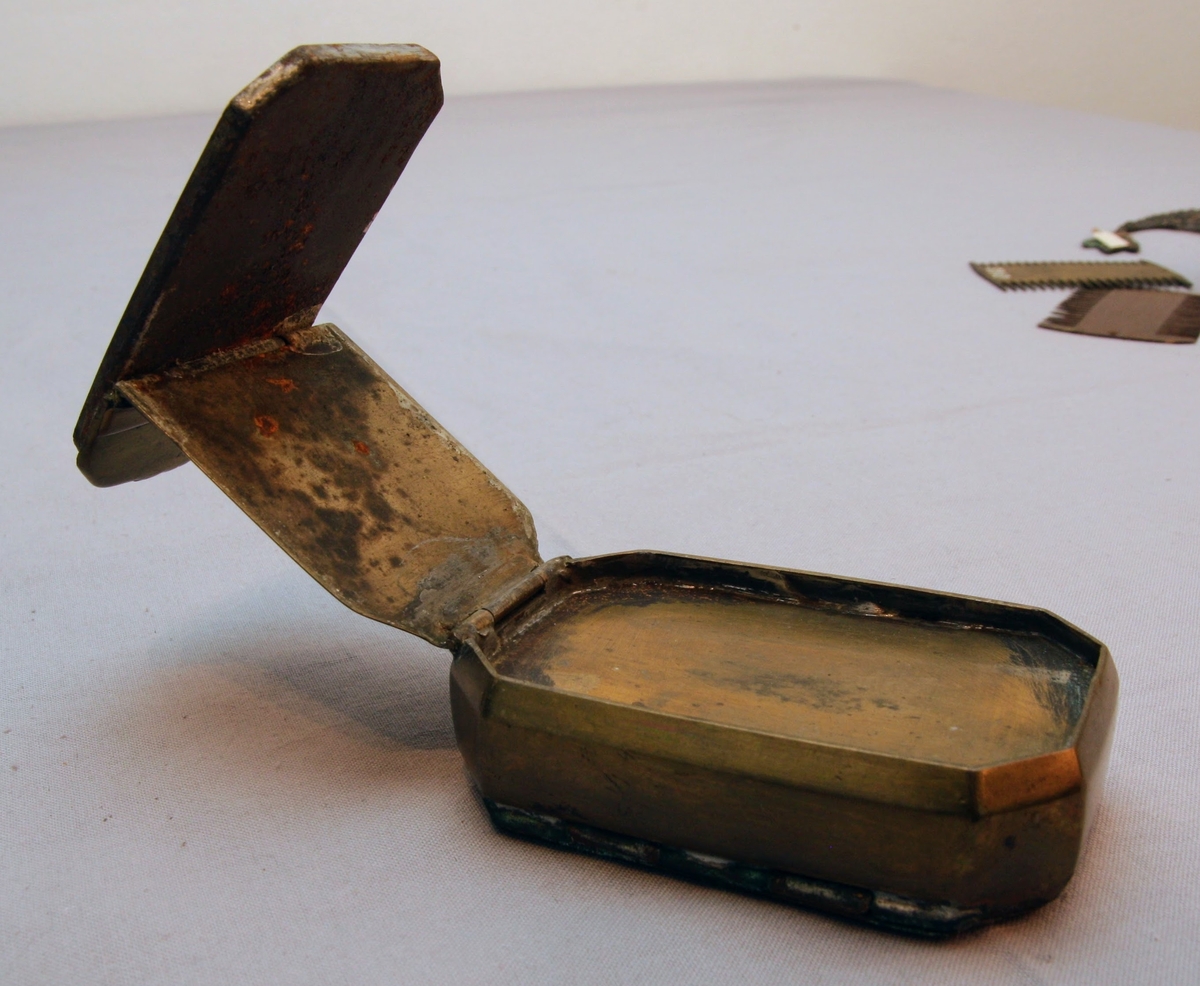 Tobaksdosa i mässing med uppfällbar spegel. Asken öppnas med ett lock i botten. Med gångjärn i ena hörnet där stödet till den uppfällbara spegeln är gängat.  Inskription i botten med bokstäverna ”APL" och siffrorna 1873 efter. Förutom inskriptionen finns inga dekorativa element på föremålet.