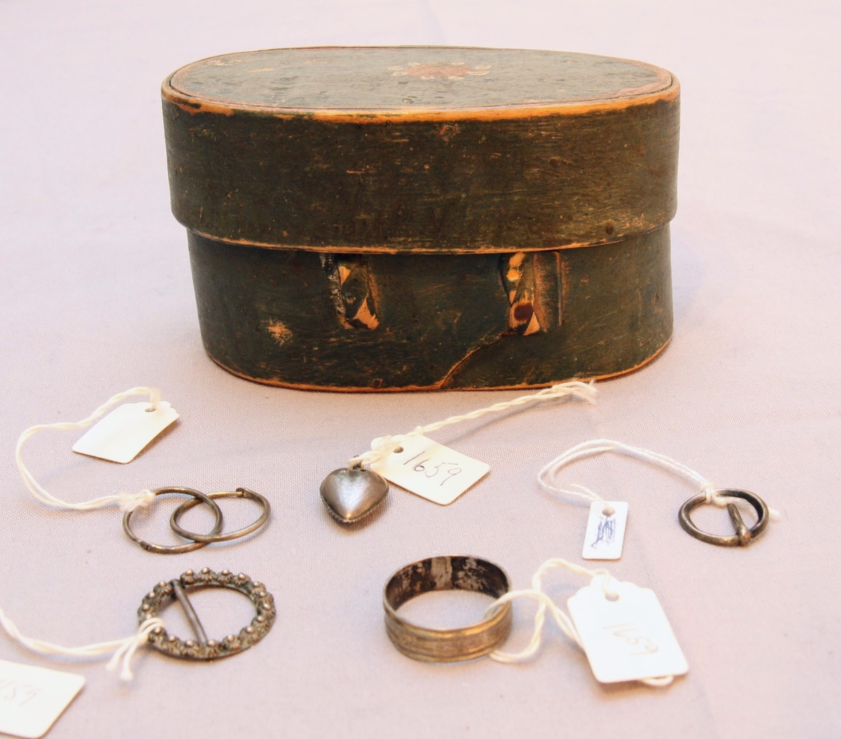 En oval ask av svept spån. Grönmålad, locket har röd kant, i mitten är en enkel ros målad. Äskan innehåller en fingerring, två mindre spännen, öronringar och ett litet hängsmycke i form av ett hjärta.