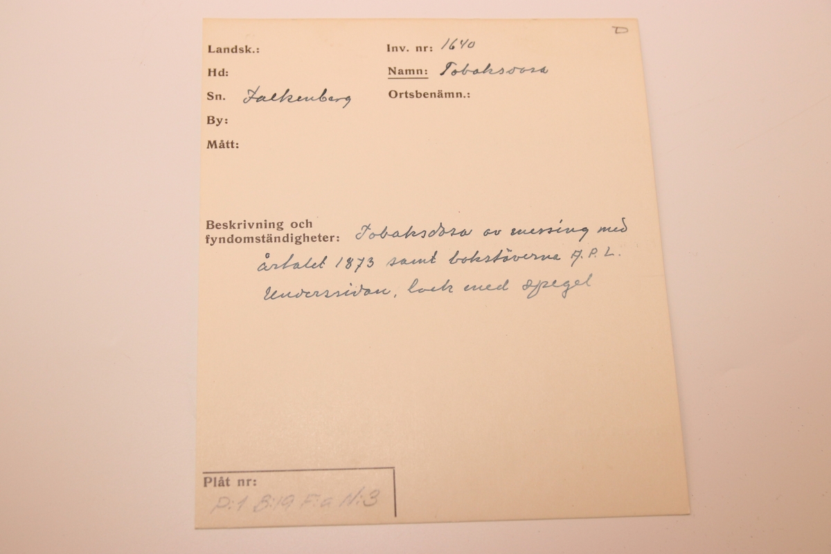Tobaksdosa i mässing med uppfällbar spegel. Asken öppnas med ett lock i botten. Med gångjärn i ena hörnet där stödet till den uppfällbara spegeln är gängat.  Inskription i botten med bokstäverna ”APL" och siffrorna 1873 efter. Förutom inskriptionen finns inga dekorativa element på föremålet.