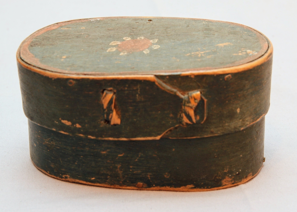 En oval ask av svept spån. Grönmålad, locket har röd kant, i mitten är en enkel ros målad. Äskan innehåller en fingerring, två mindre spännen, öronringar och ett litet hängsmycke i form av ett hjärta.