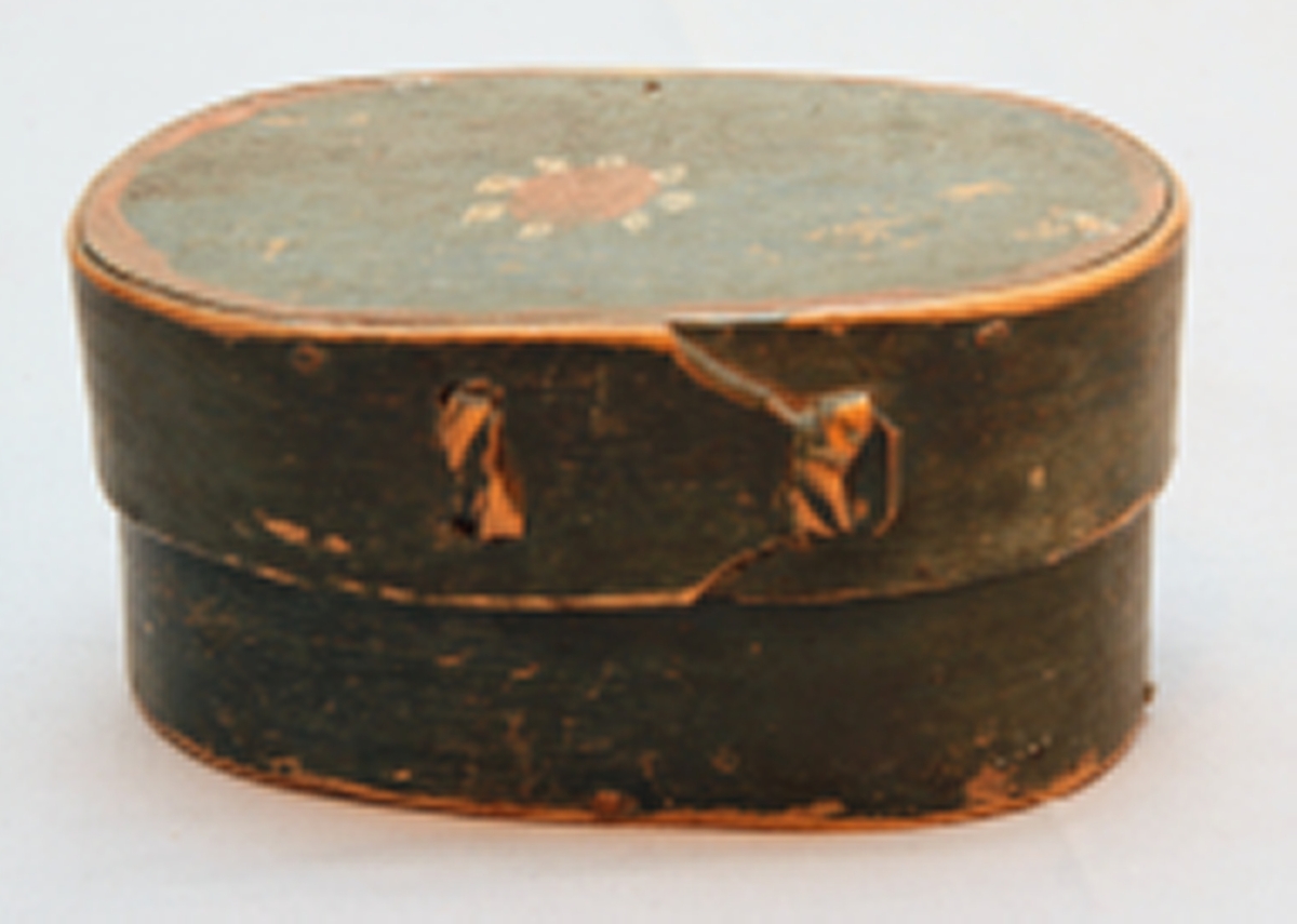 En oval ask av svept spån. Grönmålad, locket har röd kant, i mitten är en enkel ros målad. Äskan innehåller en fingerring, två mindre spännen, öronringar och ett litet hängsmycke i form av ett hjärta.