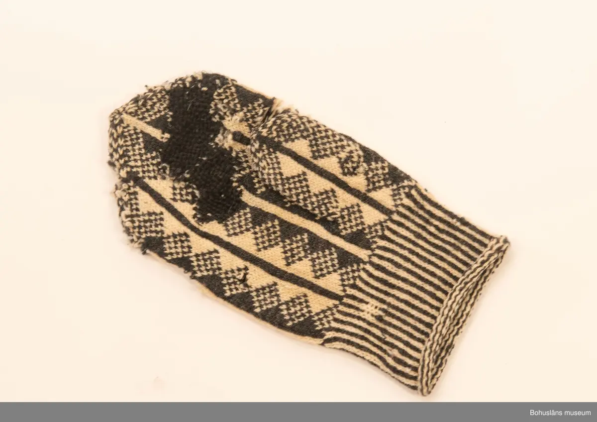 Handstickad mönstad tumvante i svart och vitt ullgarn i toppig avslutning. Slätstickad randad mönstereffekt med romber, smalrandat handledsparti. Stopplagning på insidan med svart garn.

Från gården Tun Övre prästgård, Svenneby sn.