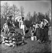 En scoutledare står vid en trave staplad ved med stenar på toppen, en fågel sitter på ett  spett över stenarna. Bredvid scoutledare står flera barbröstade pojkar, en pojke håller en död fågel i handen. En pojke skopar upp vatten från en hink till en kastrull. I bakgrunden syns träd och buskar.