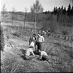 Fyra vuxna och ett barn befinner sig på en gräsplan. Två män sitter ner, en skriver med gräsmattan som underlag. I bakgrunden syns en nysådd granplantage och en ensam ungbjörk. Tre personer är påväg mot gruppen.