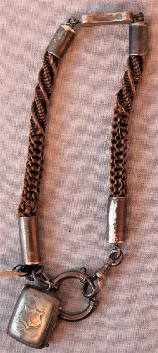 Urkedja av hår med silverbeslag. Med fyrkantig berlock med ingraverade blommor på ena sidan och en sköld på en den andra. Med en platta med ingraverat ”Minne”. - Stämpel B 7 = 1904, W. övriga stämplar oläsbara. -