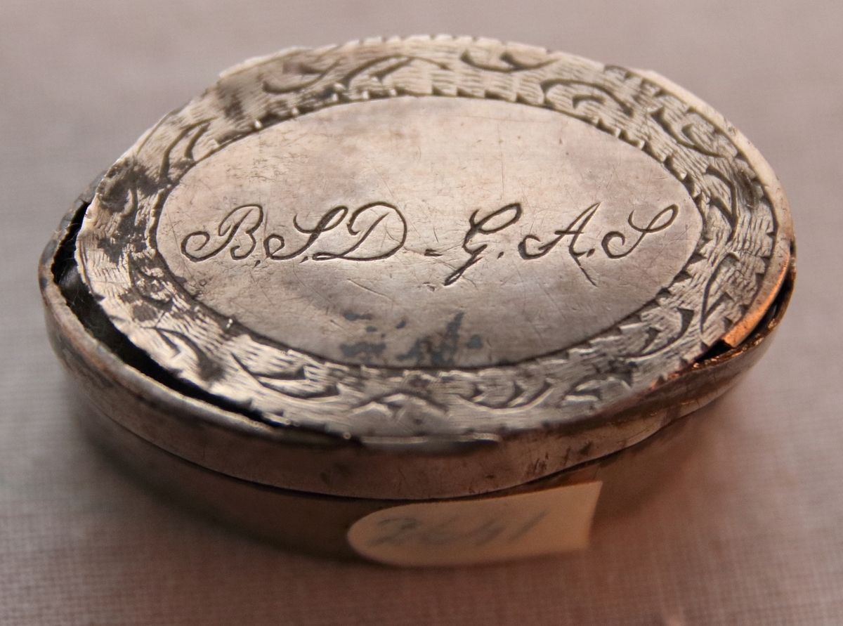 Oval dosa av silver. Med ett lock med gångjärn. Ytterkanten på ovansidan av locket har en cisilerad bård av stiliserade blad. I mitten av locket står initialerna "B. S. D. " och "G. A. S." . Asken är stämplad på insidan av botten med bokstäverna "MOB" (Michael Olof Barkman,1813-1851, Varberg) och med "N4" (år 1843) samt två svårtydda stämplar.