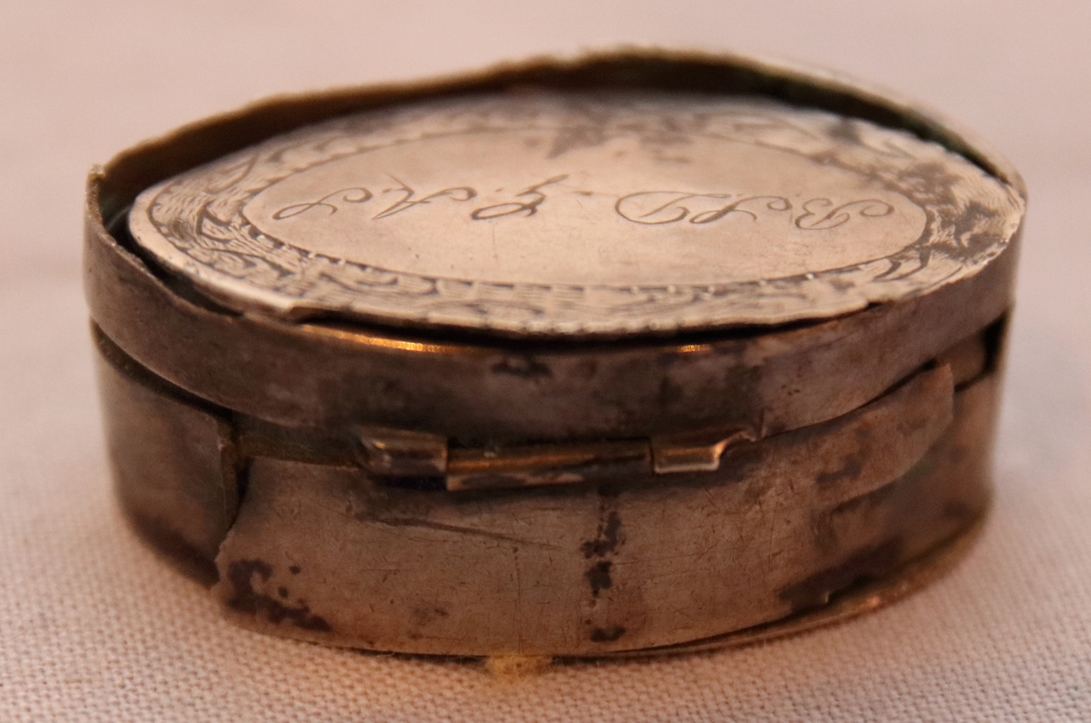 Oval dosa av silver. Med ett lock med gångjärn. Ytterkanten på ovansidan av locket har en cisilerad bård av stiliserade blad. I mitten av locket står initialerna "B. S. D. " och "G. A. S." . Asken är stämplad på insidan av botten med bokstäverna "MOB" (Michael Olof Barkman,1813-1851, Varberg) och med "N4" (år 1843) samt två svårtydda stämplar.