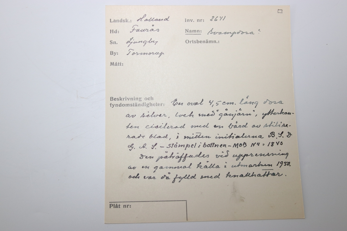 Oval dosa av silver. Med ett lock med gångjärn. Ytterkanten på ovansidan av locket har en cisilerad bård av stiliserade blad. I mitten av locket står initialerna "B. S. D. " och "G. A. S." . Asken är stämplad på insidan av botten med bokstäverna "MOB" (Michael Olof Barkman,1813-1851, Varberg) och med "N4" (år 1843) samt två svårtydda stämplar.